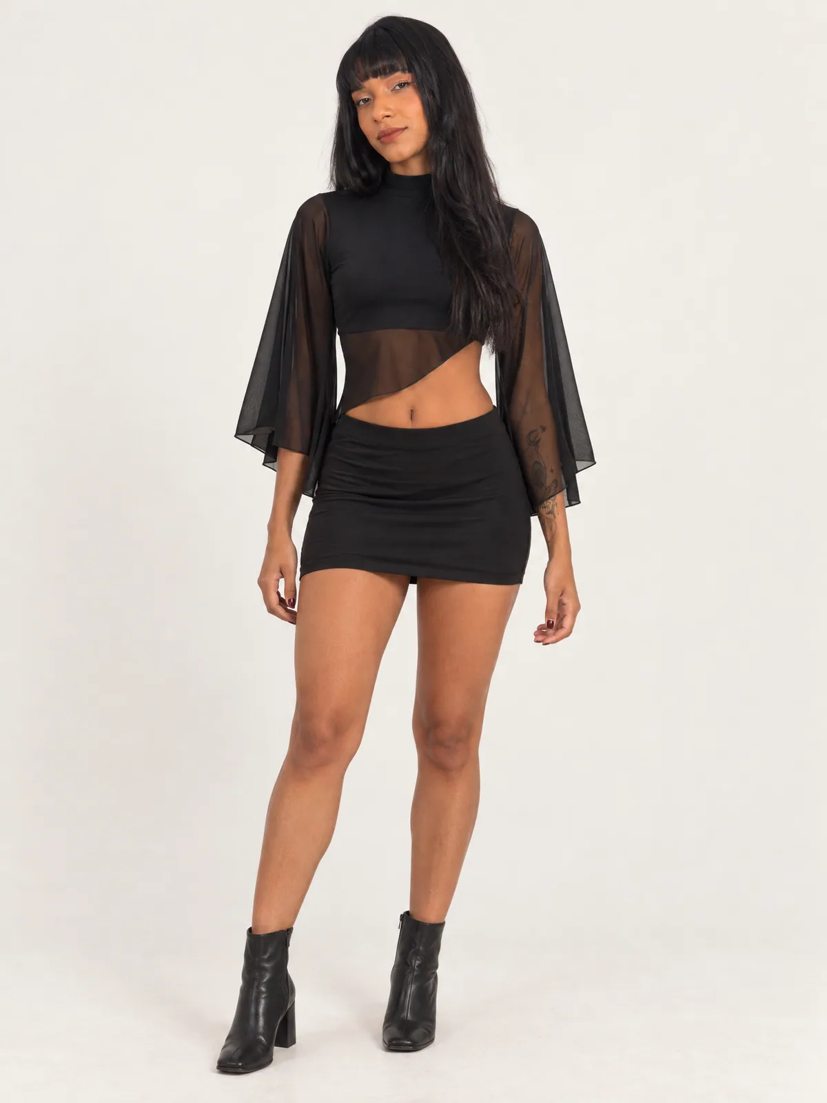 Adriely de frente com conjunto preto de gola alta, mangas capa de tule e minissaia justa